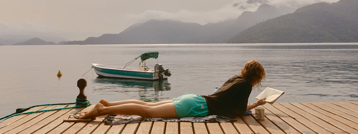 Woman reading a book on a wooden dock by a lake with mountains in the background